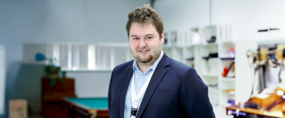 Portrait shot of a man in a dark blue suit and light blue shirt in a workshop, smiling at the camera.